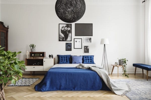 Black chandelier in navy blue bedroom in tenement house. Floor lamp between king size bed and small table with pot and clock on it. Real photo concept