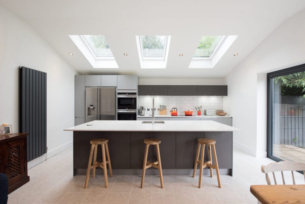 A general view of a modern fitted grey kitchen with large island, breakfast bar, white walls and sky light windows  within a home