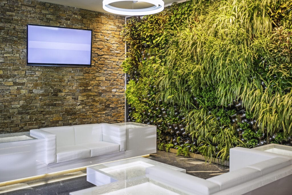 A modern designer lobby area with a flatscreen TV on a brick wall, white couches and a glass coffee table and plants growing out of a wall.