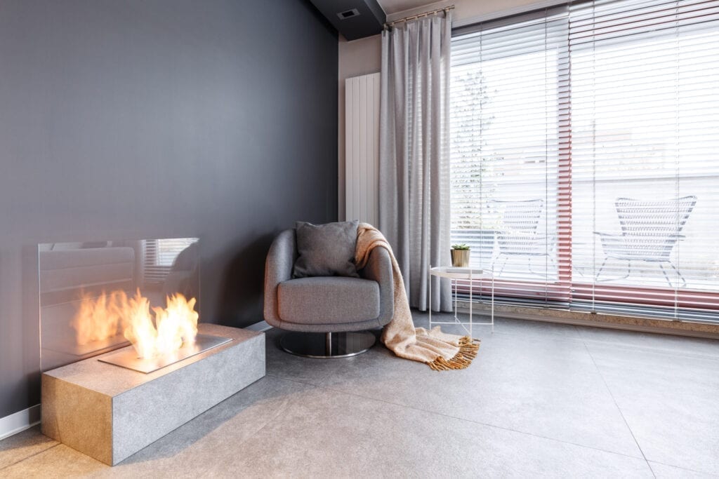 Cosy gray armchair with a pillow and warm blanket standing on marble floors in a relaxing reading corner of a bright living room interior by a lighted modern bio fireplace and big window