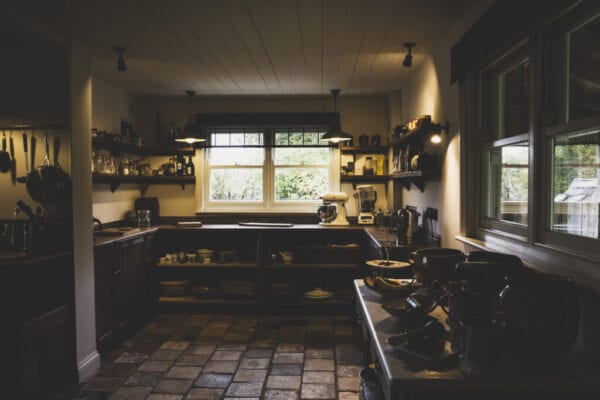 Interior view of kitchen with stone tile floor, wooden ceiling and two sash windows, antique wooden cupboards and wall shelves.