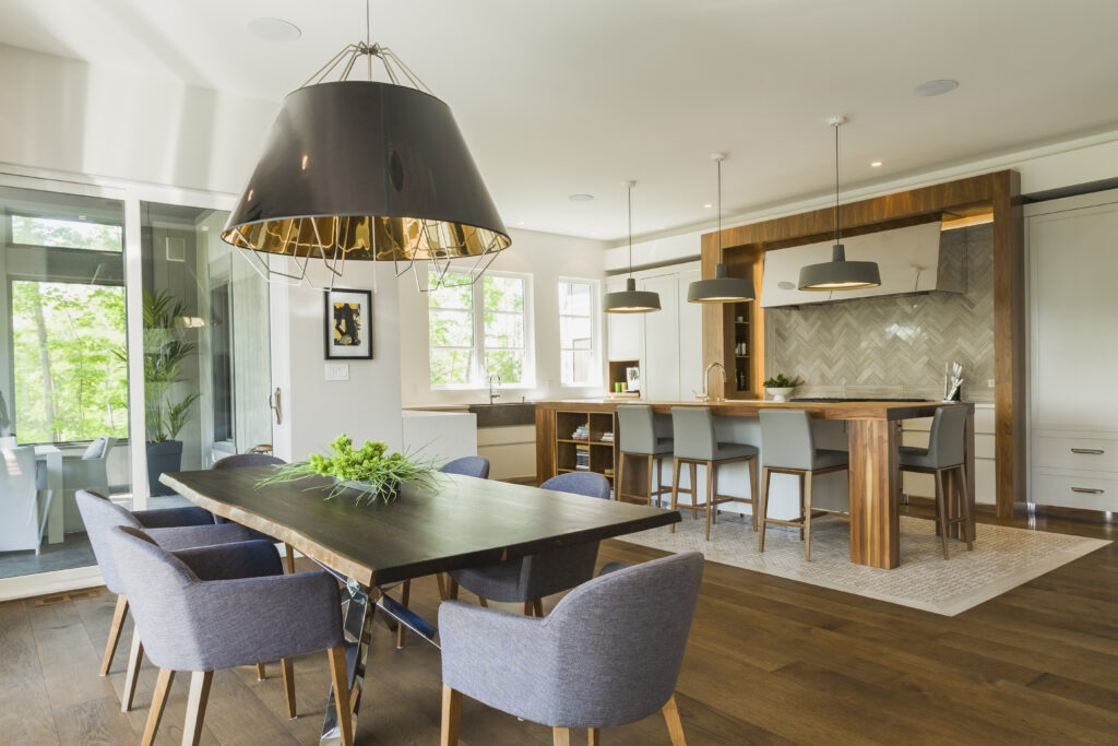 Chrome and walnut wood table with charcoal linen armchairs, large black and gold industrial style pendant lighting fixture and walnut wood island with grey leather and wooden high chairs on travertine mosaic floor surrounded by engineered hickory wood floorboards in dining room kitchen area inside a luxurious contemporary bungalow style home, Quebec, Canada. This image is property released. CUPR0327