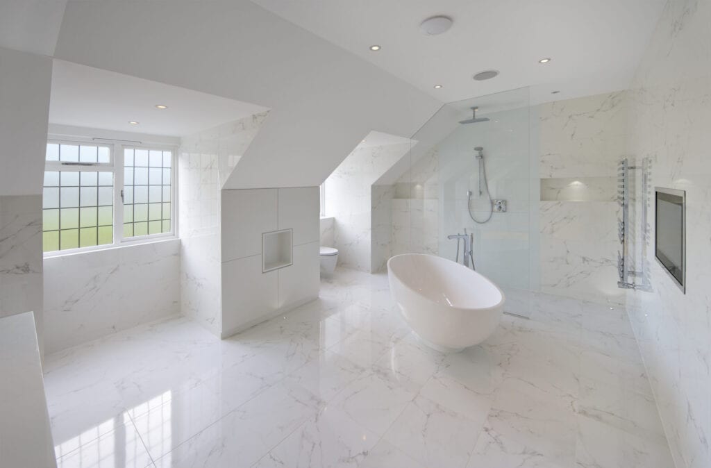 large luxury white marble lined bathroom in an expensive home with a large oval bath. In the background is the show area behind a glass screen. A hint of green, adding a bit of colour to the scene, is visible through the frosted glass windows overlooking a garden. A large TV screen has been installed flush in the wall to the right. The square feature to the left of the bath are cupboards with a recess for decorative items in the centre. more lit recesses can be seen in the shower area.