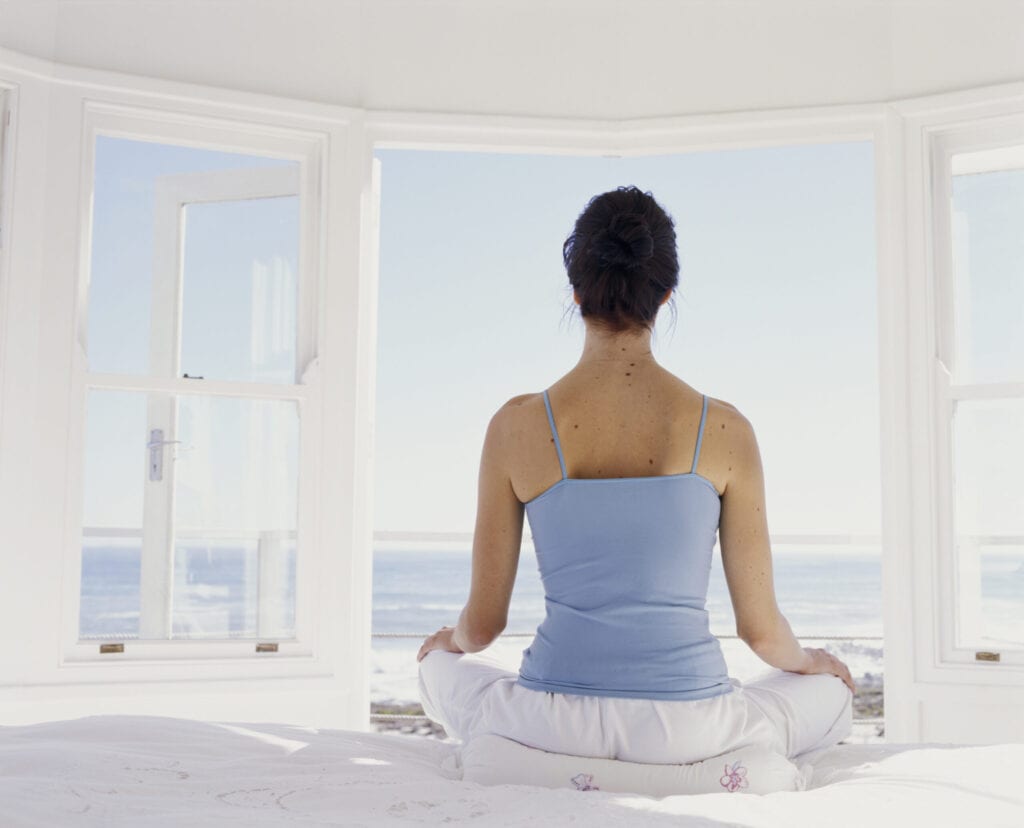 Young woman meditating on bed facing open window, rear view