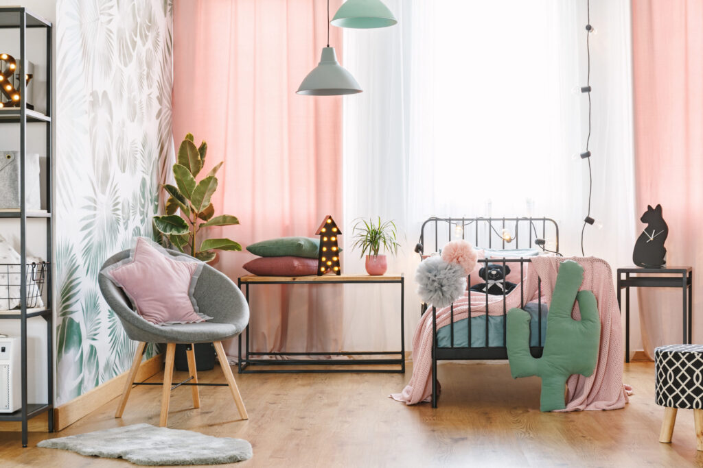 Pink pillow on grey armchair near bed in girl's bedroom interior with green cactus and lights on table