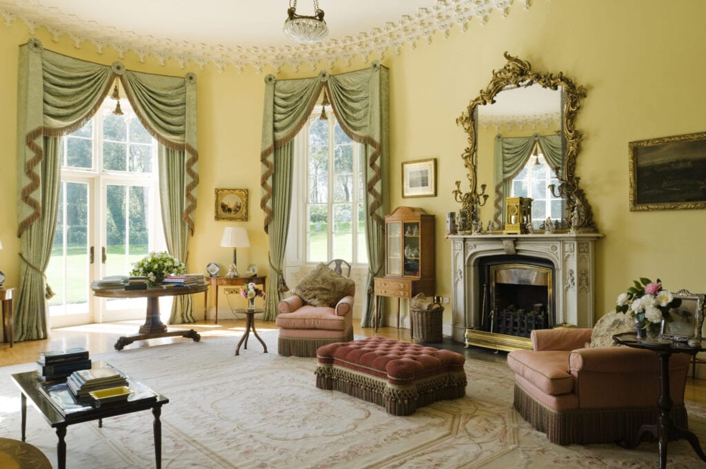Pale green fringed curtains in yellow drawing-room with gilt framed mirror, mantelpiece and red tufted ottoman