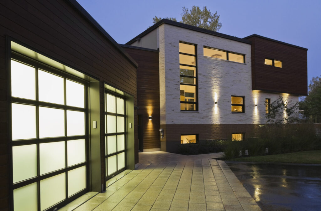 lluminated two car garage and beige stone with brown cedar wood modern cubist style residential home facade with paving stone and black asphalt driveway at dusk in summer, Quebec, Canada. This image is property released. CUPR0269