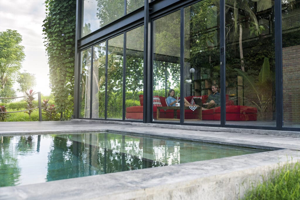 garden view through glass facade of couple with laptop and book relaxing on red couch in modern design house in front of swimming pool