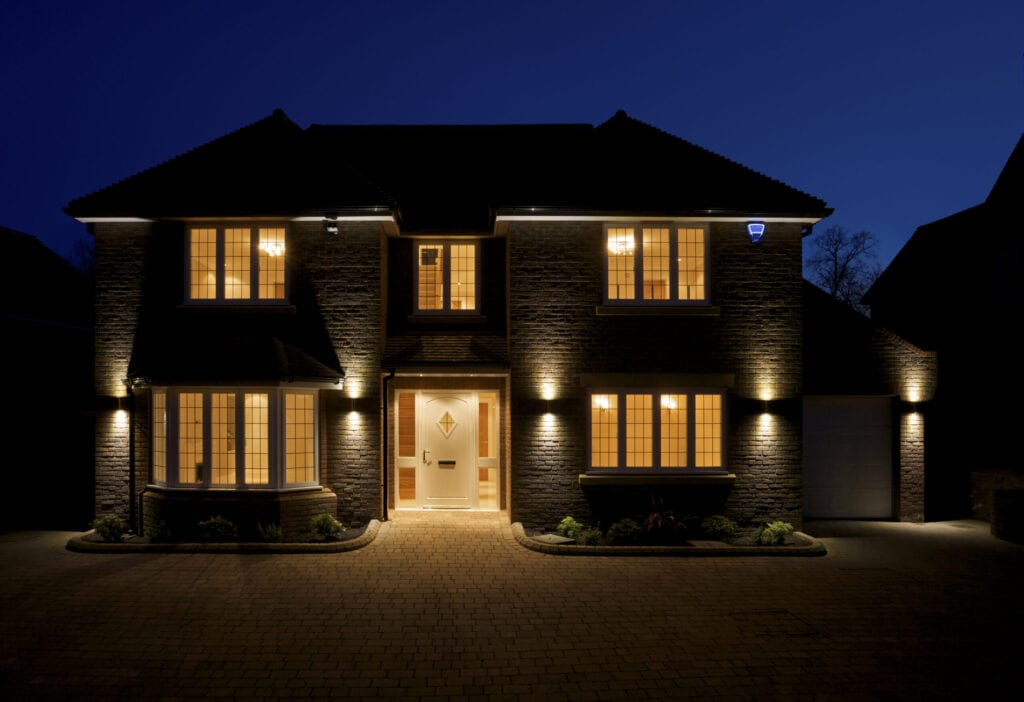 a view of the front and driveway of a lovely double fronted luxury new home, taken at dusk with all of the interior and exterior lights switched on. The branding on the alarm box has been changed to a fictional text.