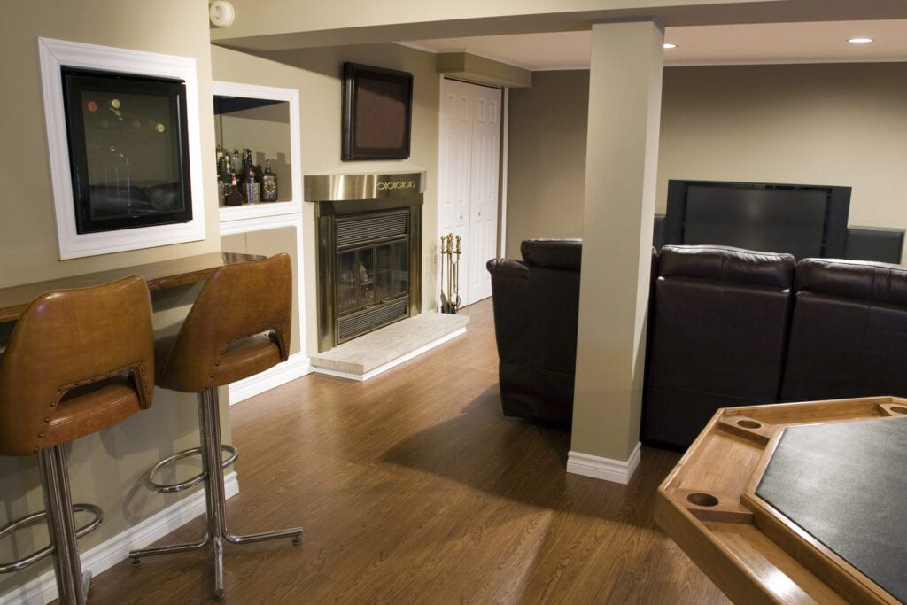 A DSLR photo of a Modern living room located in the basement of a bungalow with a wooden floor, a bar, mini fridge and poker table. A beautiful stainless fireplace close to the leather studio couch and a home cinema system.