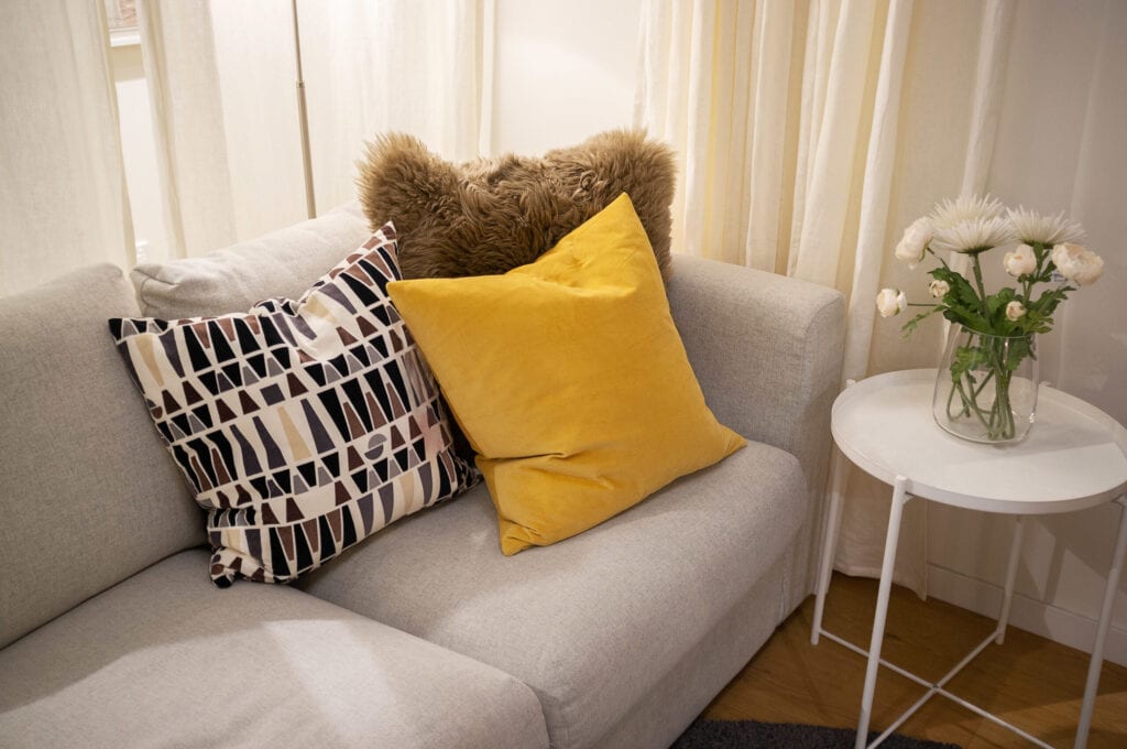 Decorative pillows on light gray textile couch with white side table and flowers - cozy interior home design