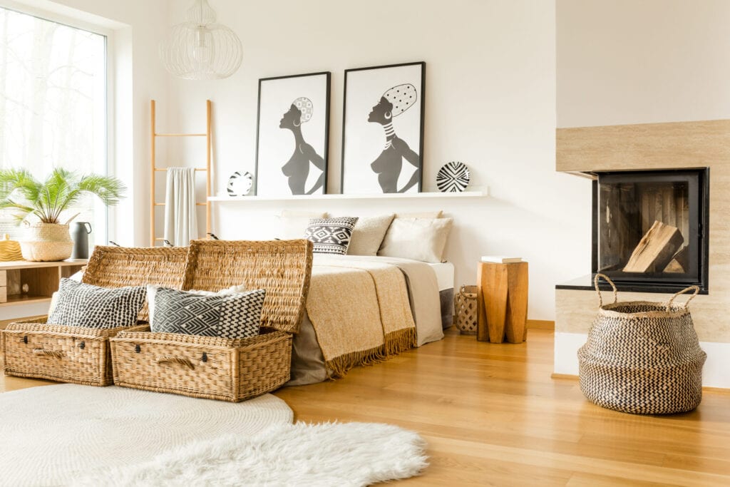 Fireplace, wattle boxes with pillows, bed, African posters in a boho bedroom interior