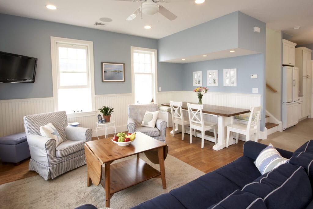 An open floor plan interior of a beach house. Living room and kitchen nook both leading into a galley kitchen and the stairwell bringing you to the three bedrooms.