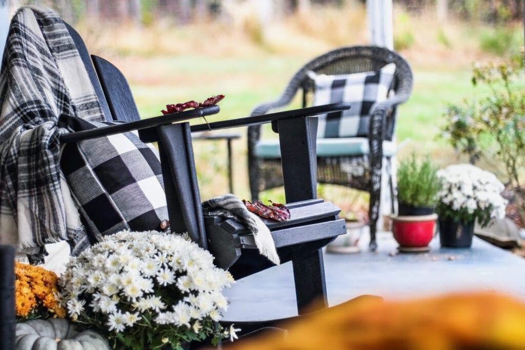 Adirondack rocking chair with traditional style buffalo check blanket and pillows on a porch or patio decorated for autumn with heirloom gourds and white and orange mums. Selective focus with garden blurred in the background.