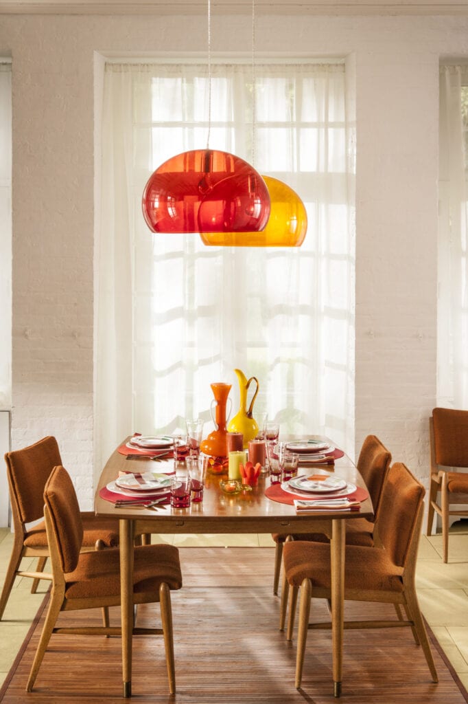 A women setting a colorful table in a bright Midcentury Modern dining room,with orange accessories