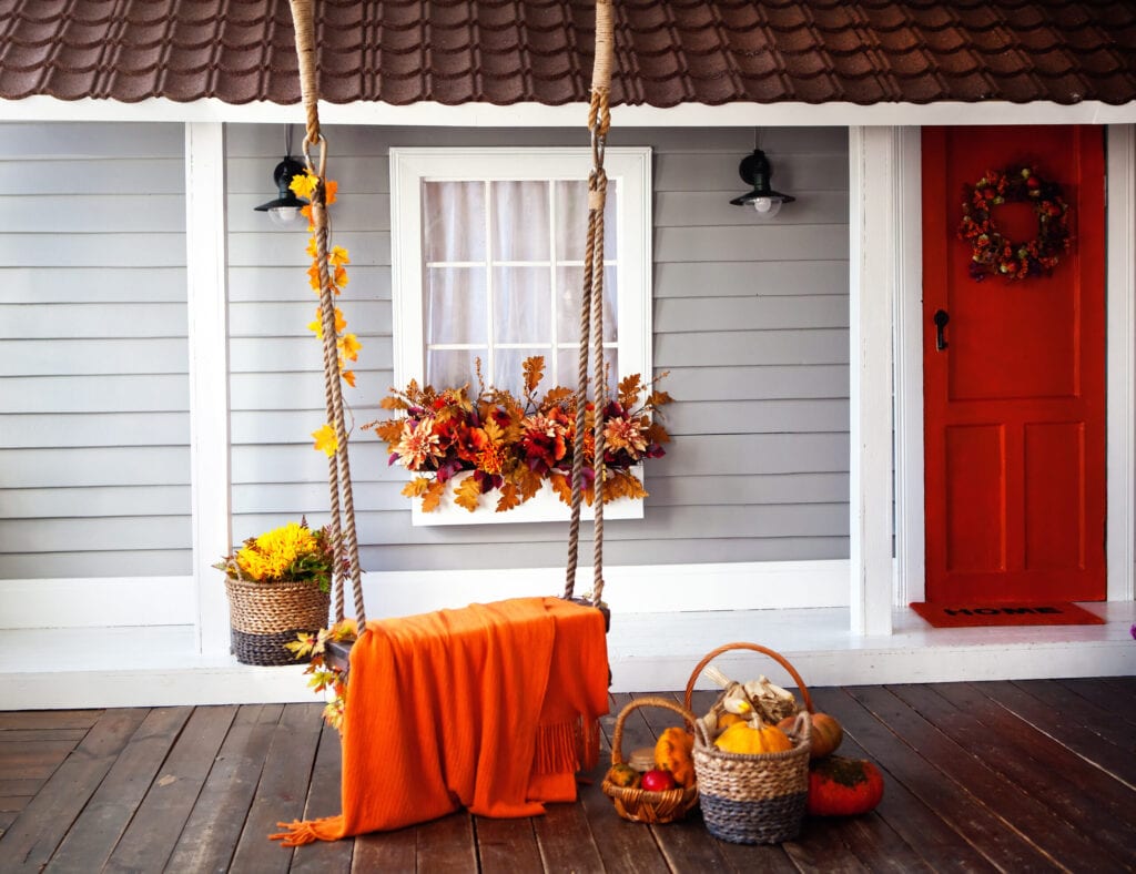 interior of an autumn patio. swing is adorned with autumn leaves and orange knitted plaid. Basket with pumpkins and autumn vegetables. The window is decorated with autumn decor. Bord door.
