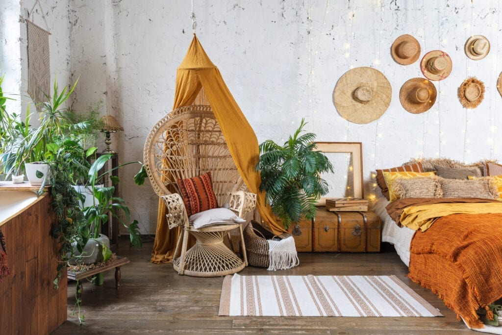 Elegant and quiet bohemian room with cozy interior, wicker chair, pillows, cushions, green plants in flower pot, bed and rug on wooden floor