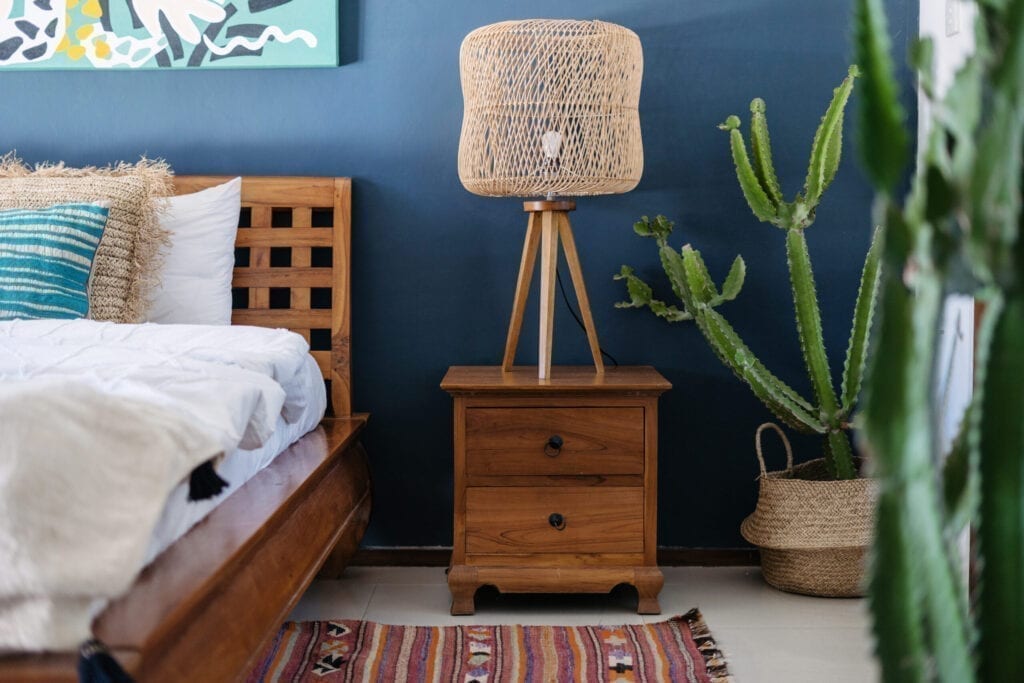 Vertical photo of lamp on wooden bedside table, ethnic decor, comfortable bed, woven ornament carpet and exotical cactus plant in basket. Concept of elegant bedroom interior in cozy house