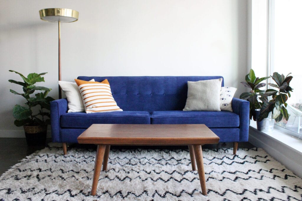 Low angle shot of a royal blue suede mid century modern 4 seater sofa and teak coffee table. Set in a modern apartment living room complete with mid century furnishings, moroccan rug and potted plants.