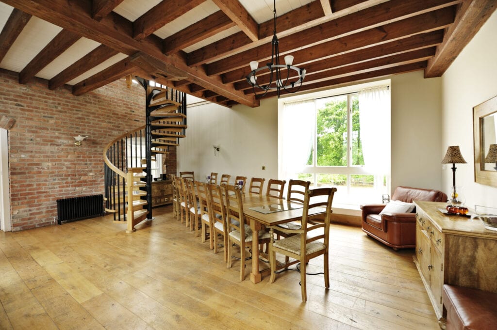 Luxury dining room with table and 14 chairs, open spiral staircase heading to upper storey, feature brick wall and sofa corner, in renovated old house. View to garden through window"