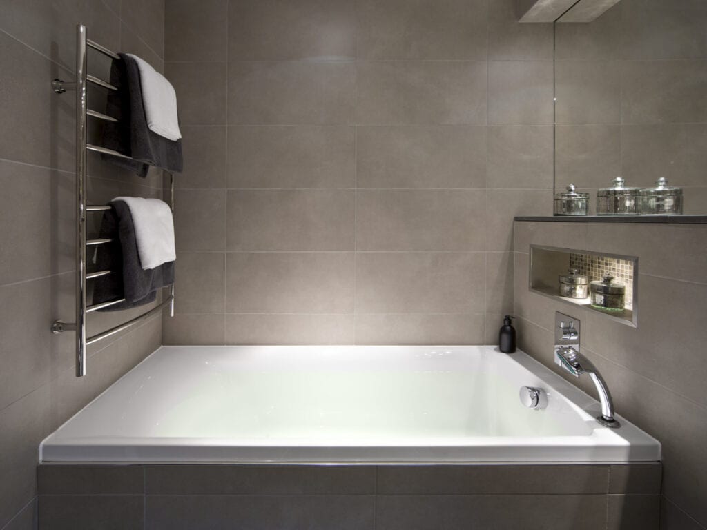 a nice steaming-hot bath ready and waiting... A large modern bath tub full of water, surrounded by fine bronze coloured marble tiles. To the left, a towel rail holds brown and white towels. On the right, metalised glass receptacles sit decoratively in an elegant recess and also on a shelf below the mirror. A brown soap dispenser sits in the corner, opposite a chrome hand shower.