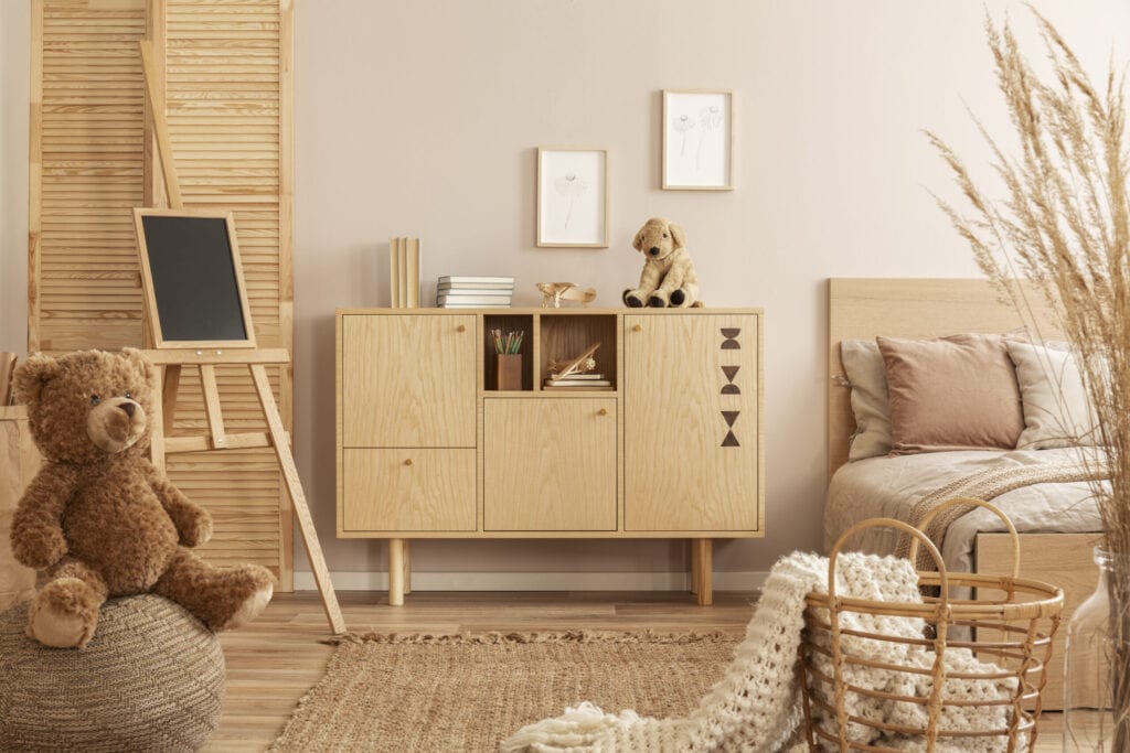 Teddy bear on pouf and small blackboard on easel in stylish kid's bedroom with wooden cabinet and beige bedding on bed