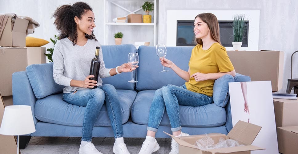 Two young women sit on their couch drinking wine as they say goodbye to their old house.