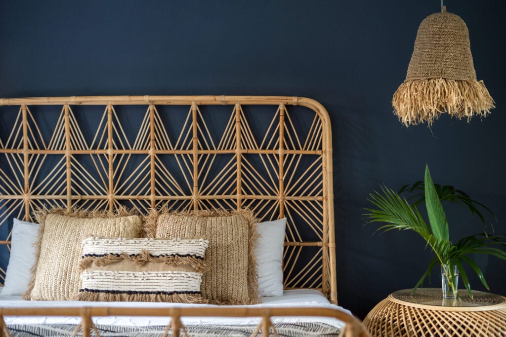 Traditional asian interior bedroom at cozy house with ethnic decor, wicker headrest at comfortable bamboo bed, lamp over nightstand table and natural green plant composition