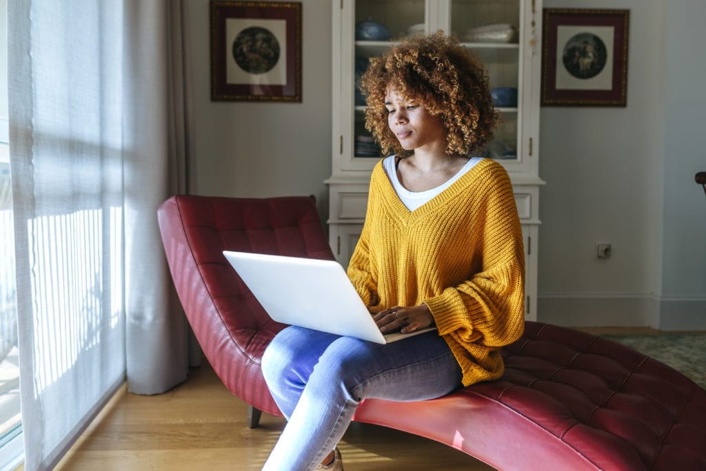 Young woman sitting on chaise lounge at home using laptop