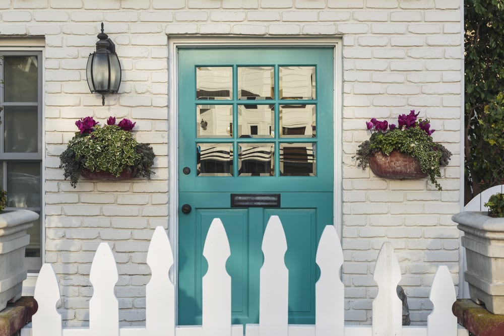 A teal wooden front door to a home, with white picket fence gate in foreground. The door is framed by two flower planters, and detail of the white, brick house.