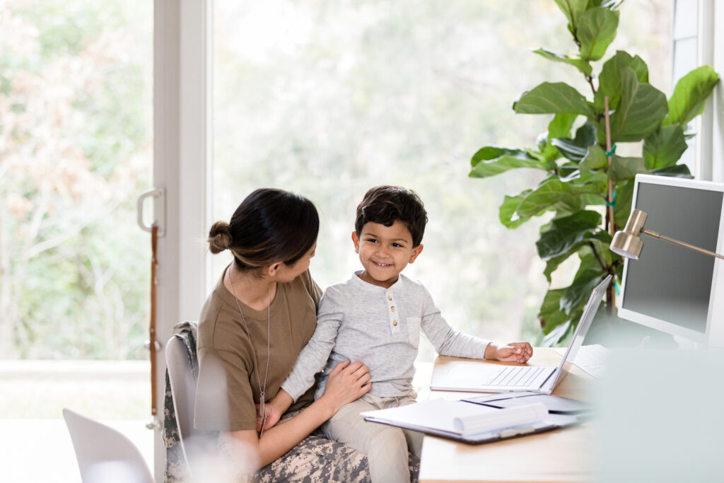 The young boy smiles since he gets to sit in his soldier mother's lap as she works at her desk.