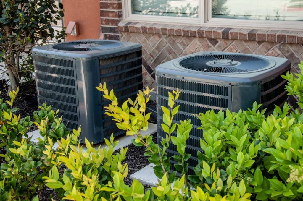 Heating and air conditioning units on the side of a brick building, hidden by shrubs