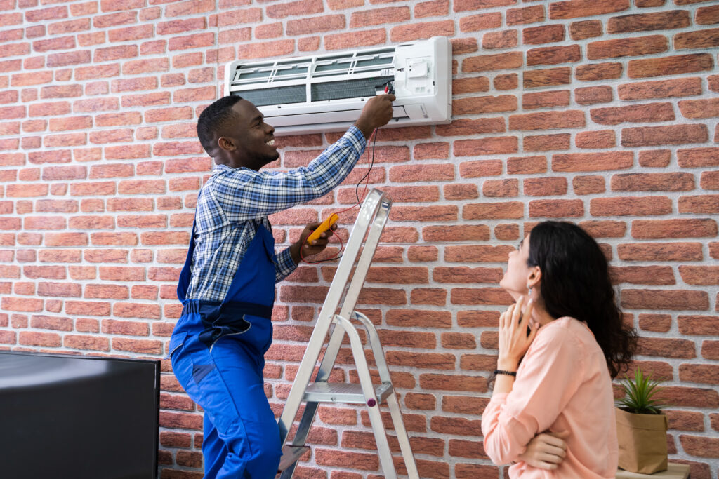 Young Woman Looking At Male Technician Repairing Air Conditioner Mounted On Brick Wall