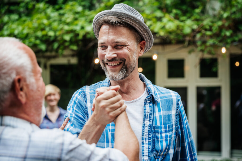 An elderly man shaking hands with one of his family members before sitting down for a barbecue meal together.