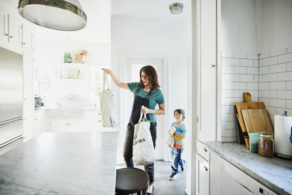 Pregnant mother bringing groceries in canvas bags into kitchen with young son