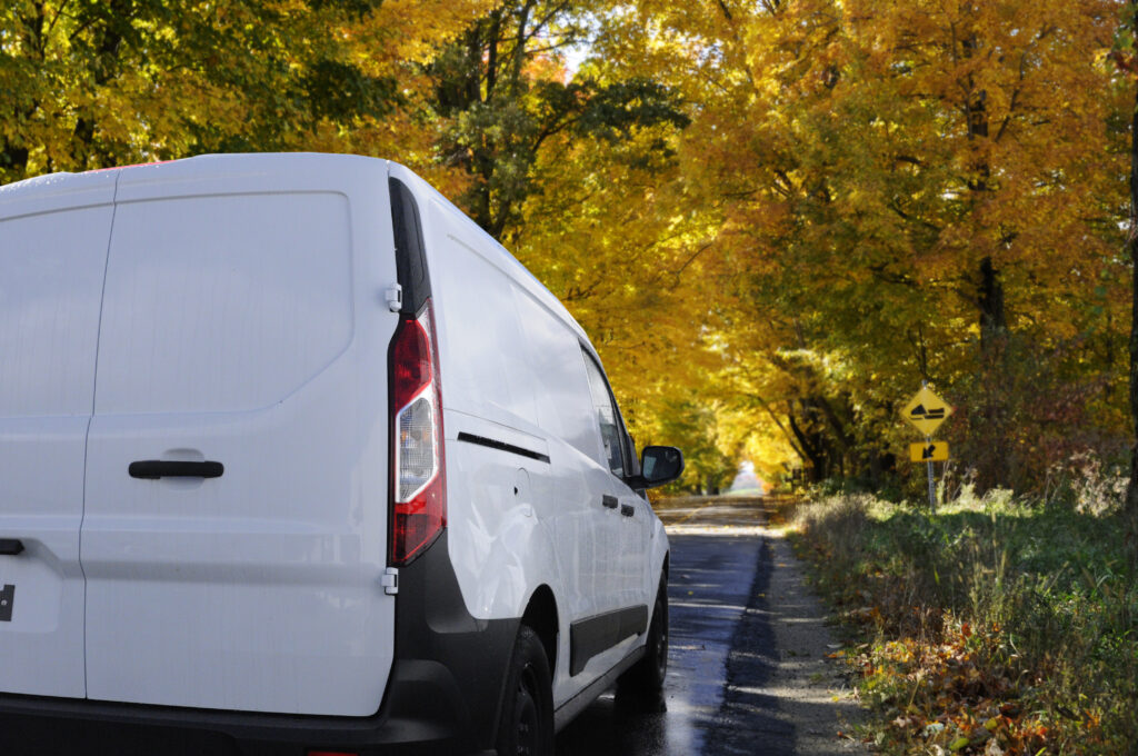 White delivery van on a rural road at the peak of autumn crossing a colorful tree tunnel