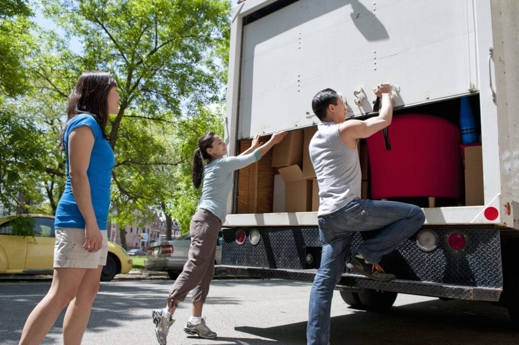 group of friends standing at the back of small moving truck, lifting up rear door, truck filled with moving boxes and furniture, urban environment