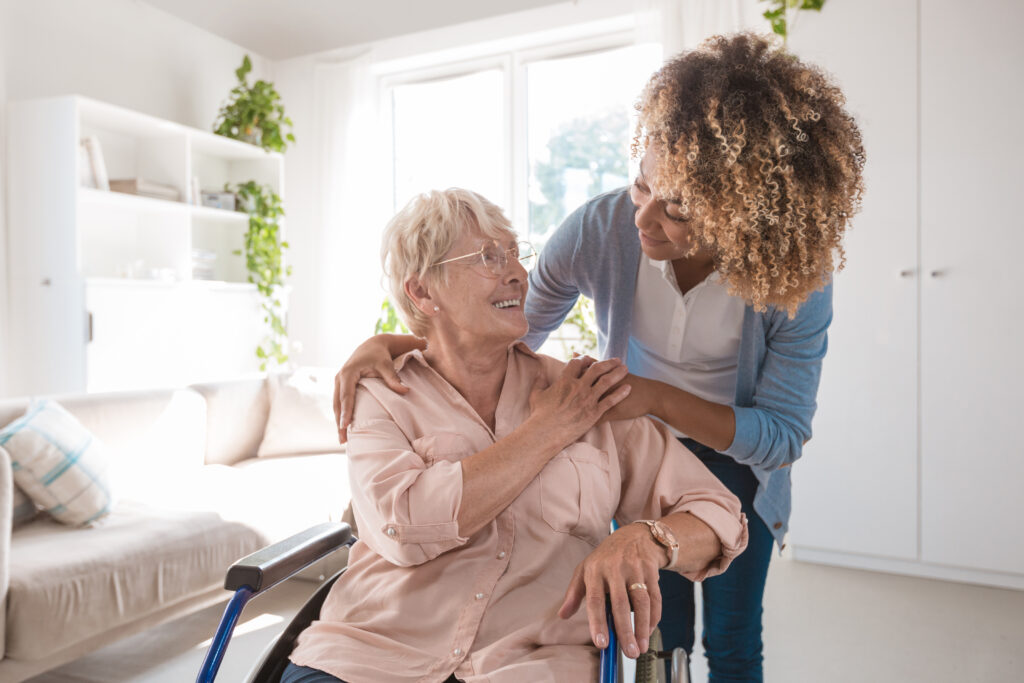 Senior woman sitting in wheelchair and talking with afro american home caregiver, smiling to her.