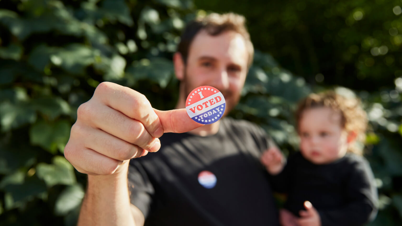 A father standing outside holding his baby, with an "I Voted Sticker" on his thumb