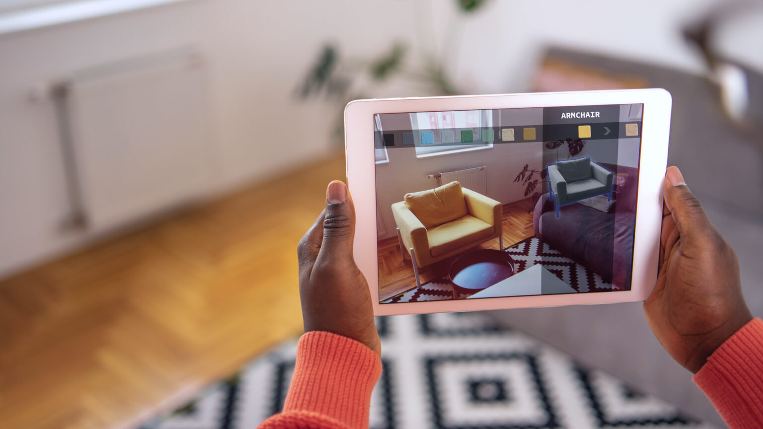 Hands hold a digital tablet with the augmented reality app, which shows the armchair in the living room. In the app, you can choose the model of a chair and the color and texture of the fabric.