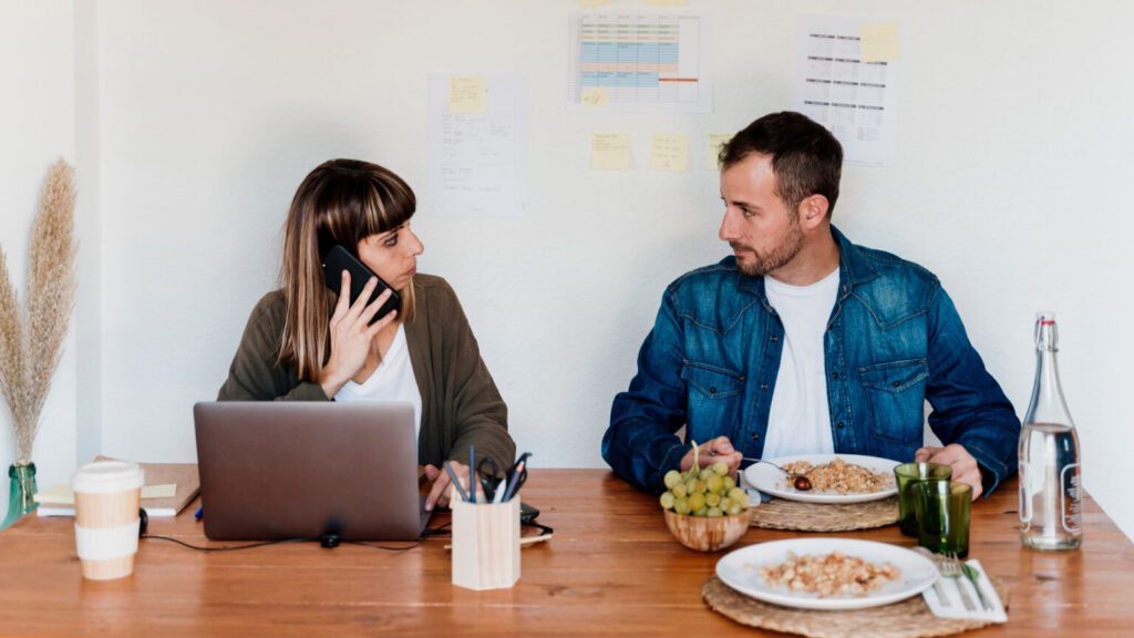 Businessman having food while female colleague talking on mobile phone at home office