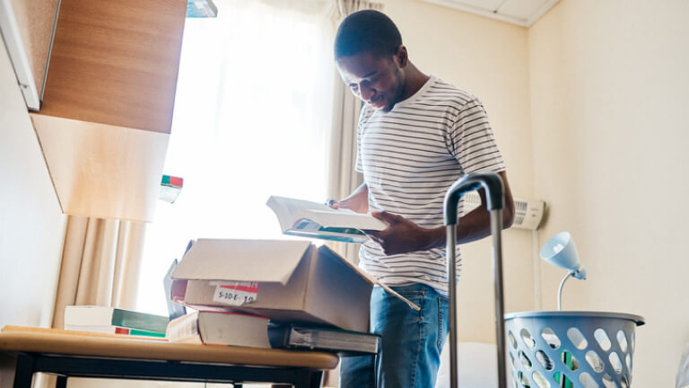 Young man looking at page in book standing in his dorm room