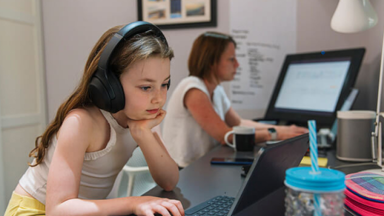 Young girl remote schooling in study. Mother is working in the background