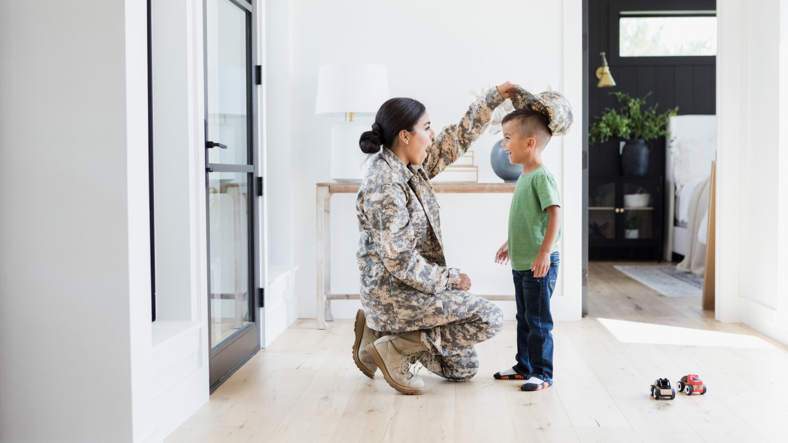 Little boy wears mom's military hat in the entryway of home.