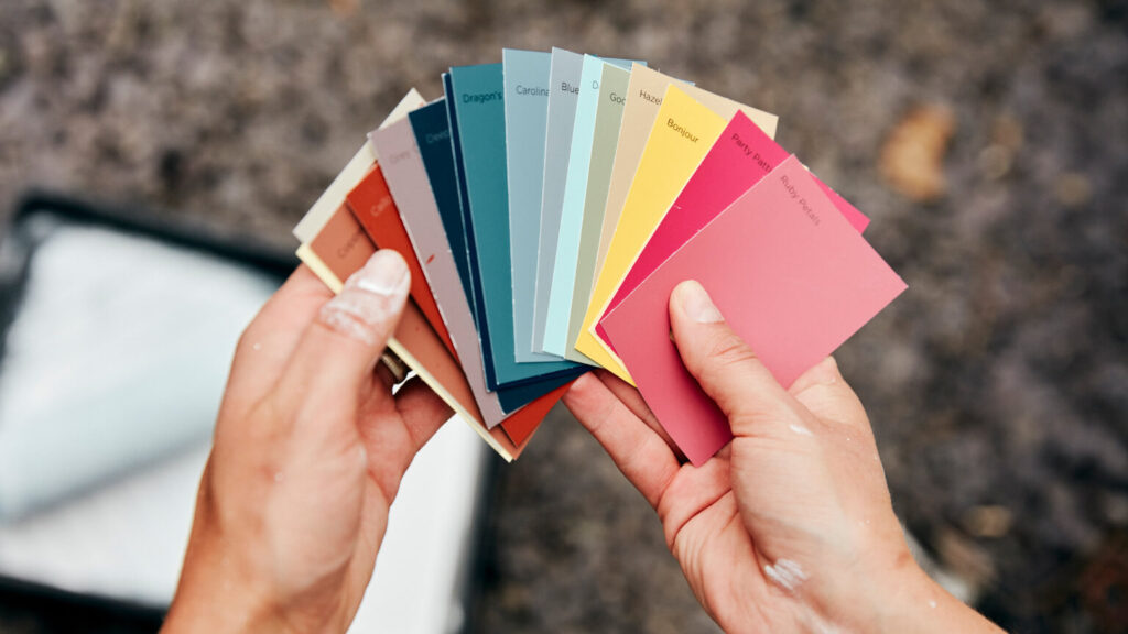 A person holding a paint color chart, and a paint tray and roller in the background.