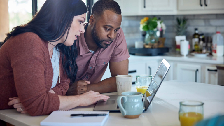 Caucasian woman and African American man sit at kitchen counter with breakfast working with pen, paper and laptop.