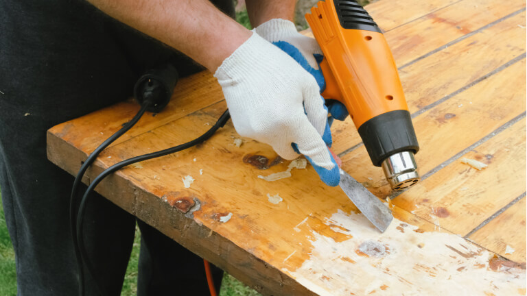 Man removing old varnish from wood using scraper and heat gun.