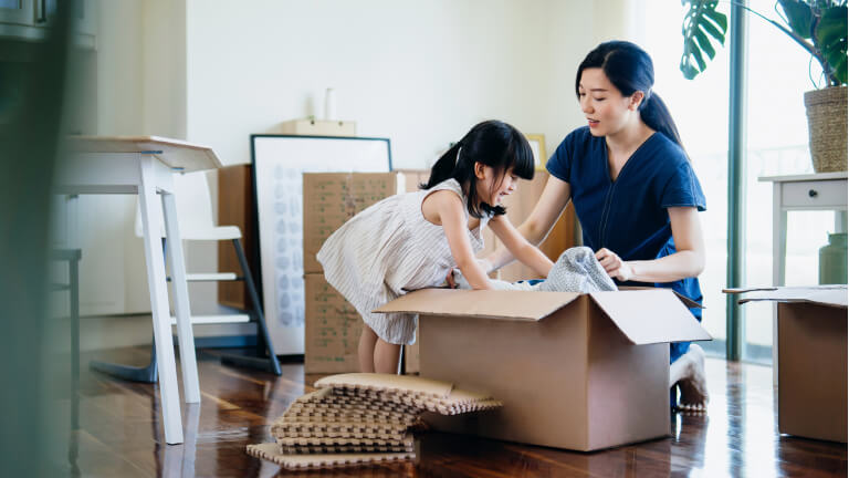 Young Asian family moving into a new apartment. Lovely little daughter helping her mother to pack / unpack belongings into cardboard box in the living room. Home moving, migration, relocation concept