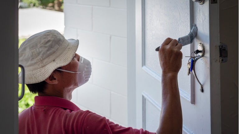 Man wearing facemask, painting a white door with paintbrush.